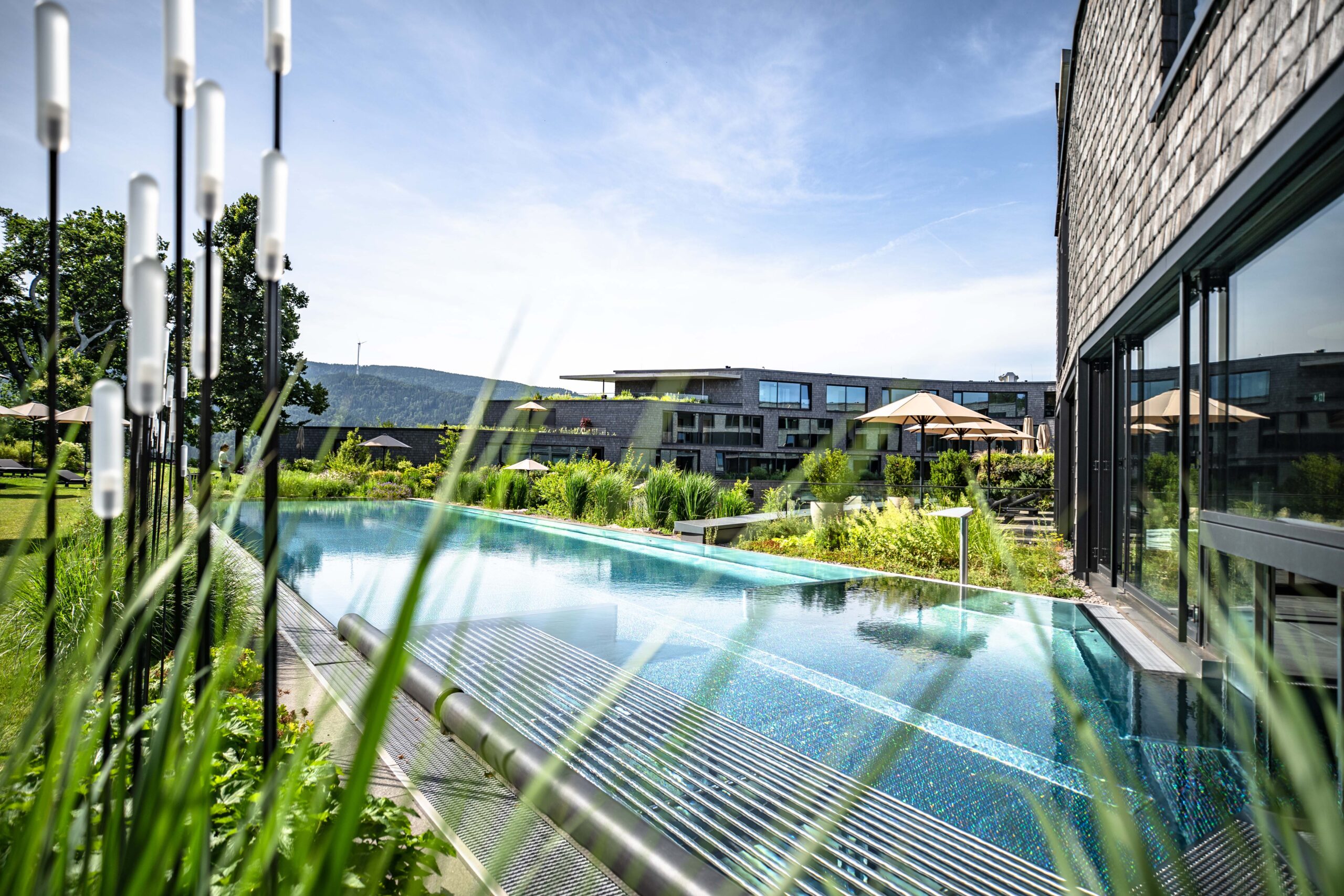 Ein modernes Freibad neben einem modernen Gebäude, umgeben von Grünpflanzen und Sonnenschirmen, lädt Sie zu purer GenussZeit ein, mit den Bergen im Hintergrund unter blauem Himmel.