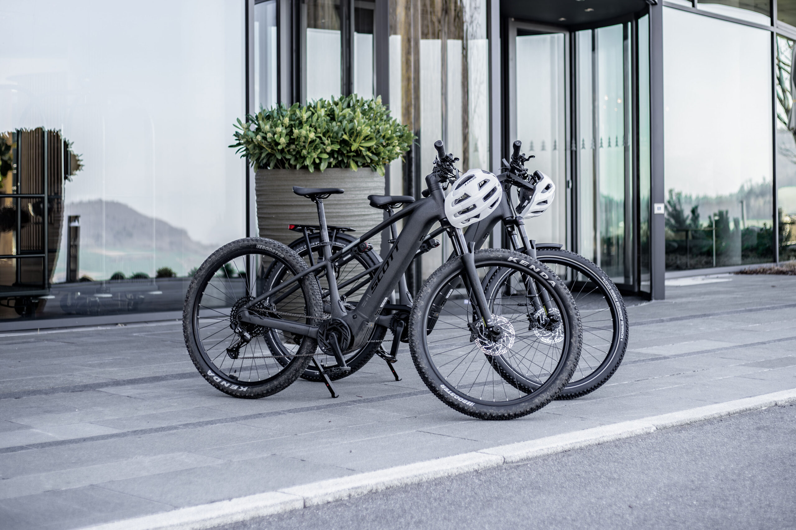 Zwei schwarze E-Bikes mit weißen Helmen stehen nebeneinander auf einem grauen Steinpflaster vor dem Eingang eines modernen Glasgebäudes, in dessen Nähe eine große Topfpflanze steht.