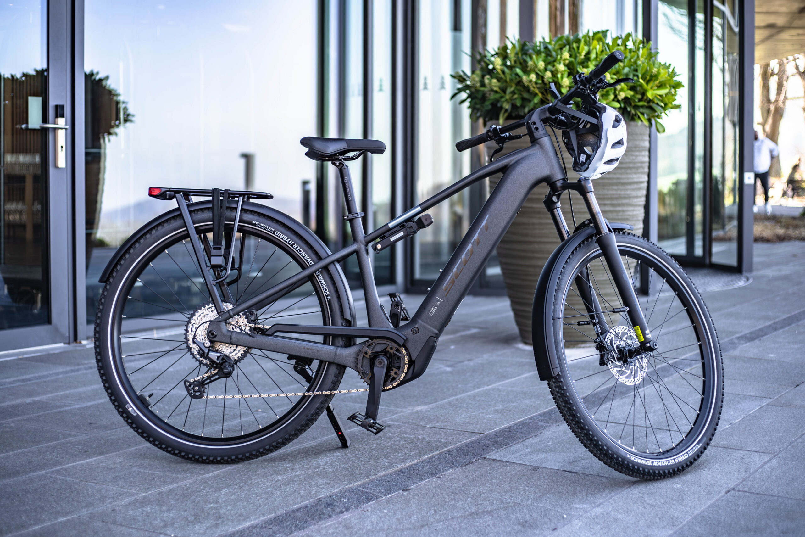 A black electric bicycle with a rear cargo rack and a white helmet hanging from the handlebars is parked on a tiled sidewalk outside a modern glass building with large potted plants.