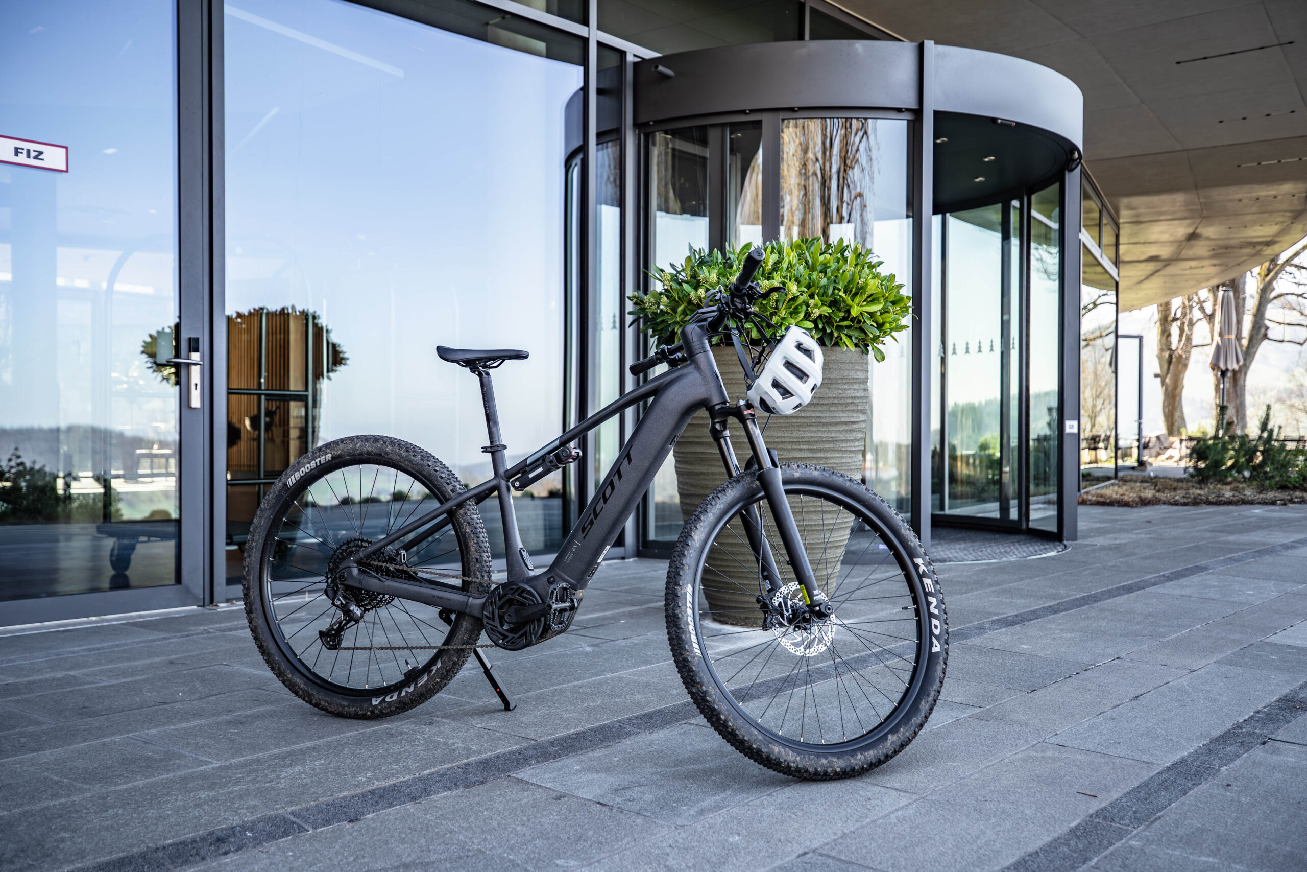 A black mountain bike with a white helmet on the handlebars is parked outside a modern building with large glass windows and a revolving door. Green plants are visible near the entrance.
