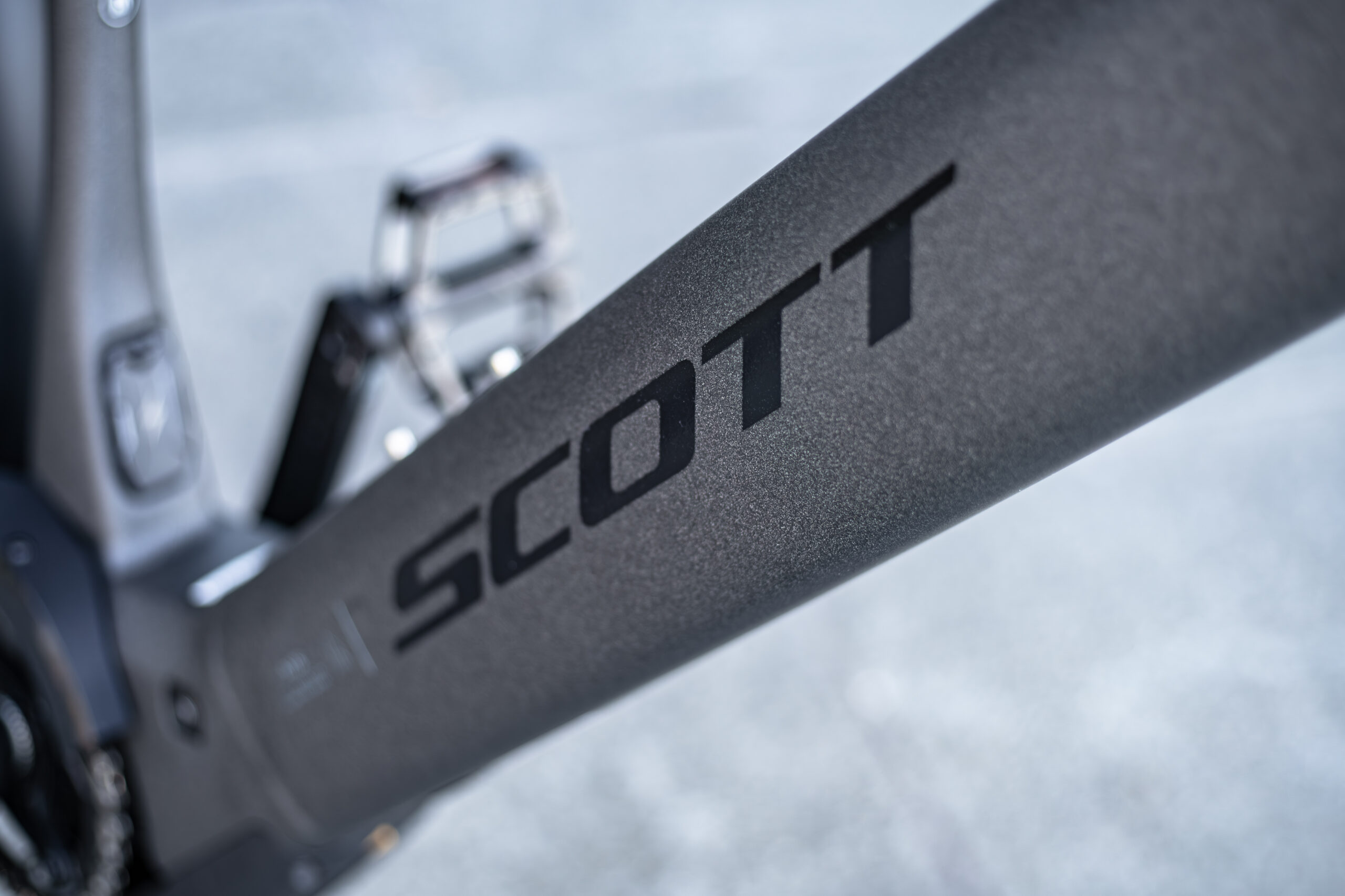 Close-up of a bicycle frame with the brand name SCOTT printed in bold black letters. A bike pedal and part of the gear mechanism are visible in the background.
