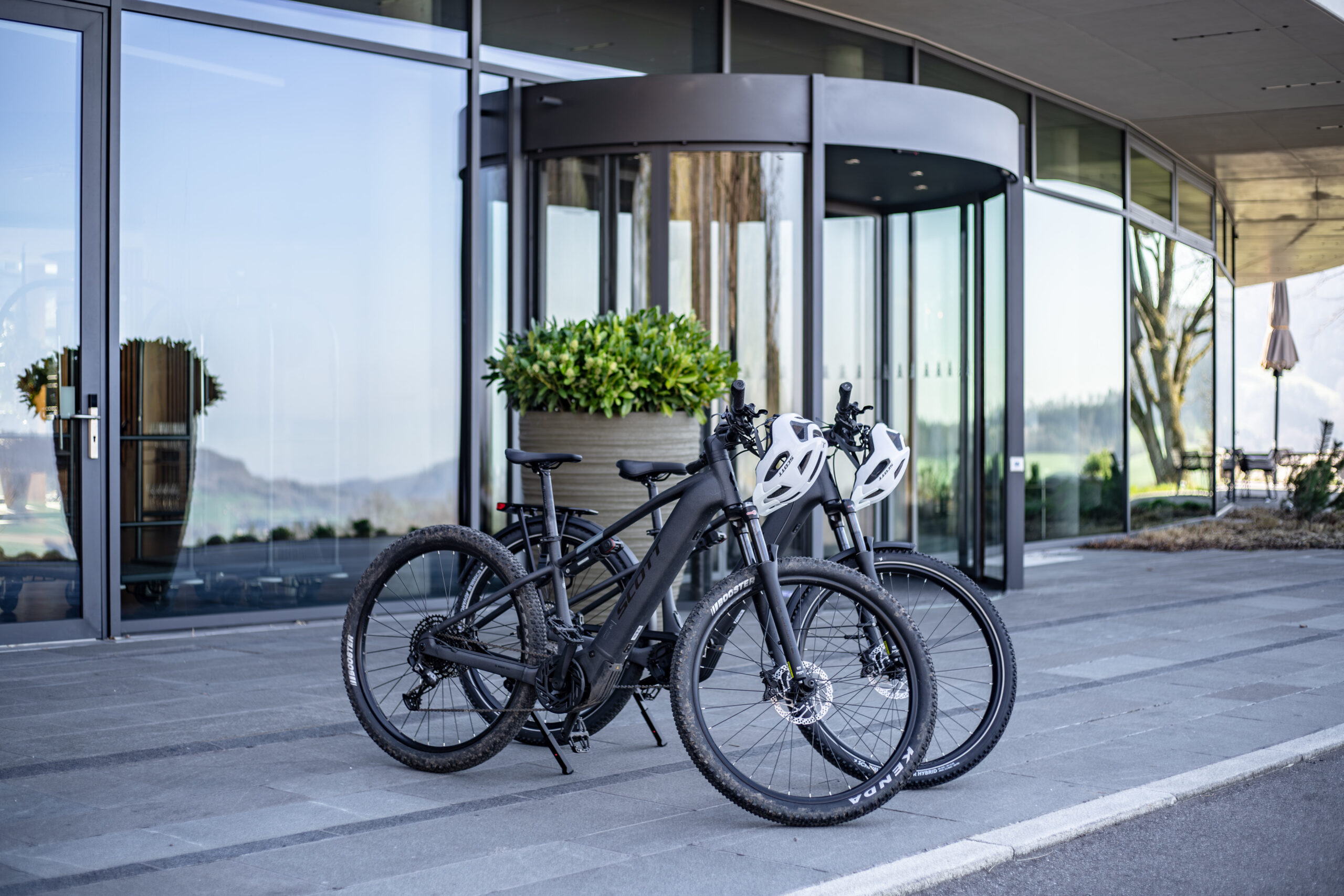 Two black mountain bikes with white helmets on the handlebars are parked on a sidewalk in front of a modern glass building with a revolving door and large potted plants.