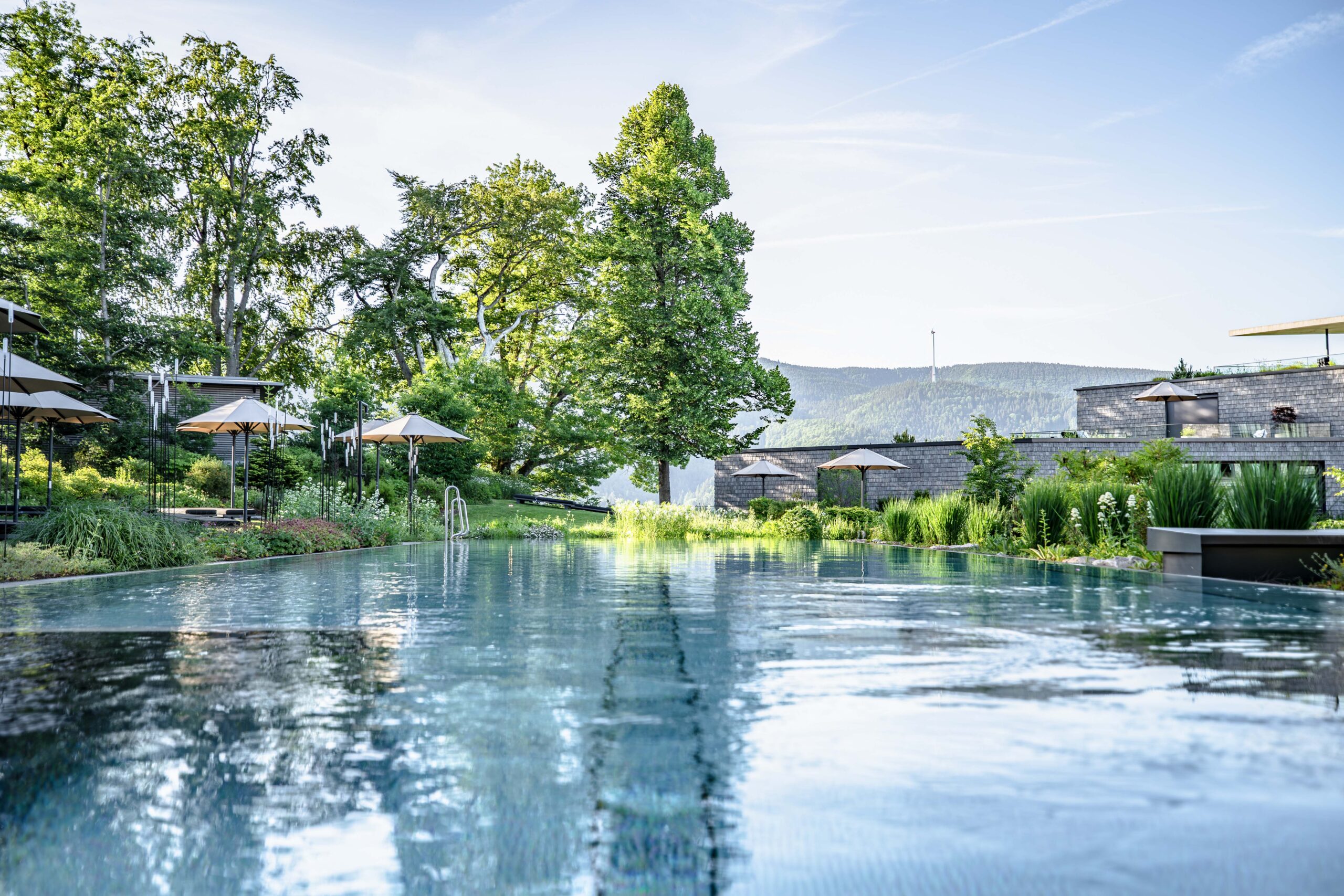 A serene outdoor infinity pool surrounded by lush greenery, lounge chairs with umbrellas, and modern buildings, with mountains visible in the background under a clear sky.