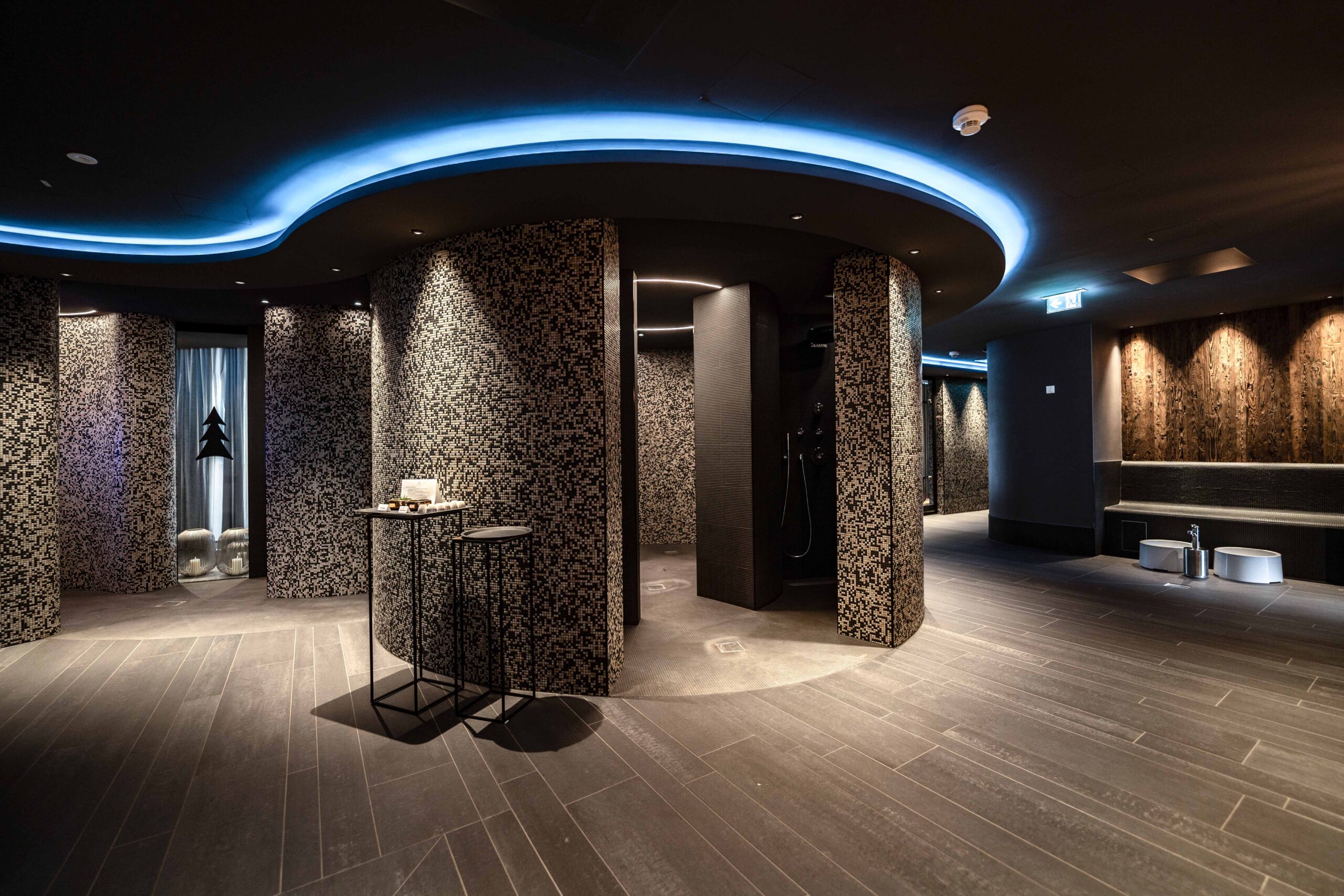 Modernes Spa-Interieur mit geschwungenen, strukturierten Wänden, sanfter Beleuchtung und einem runden Deckenakzent mit blauen Lichtern. In dem gedämpft beleuchteten, eleganten Raum sind kleine Tische und Bänke verteilt.