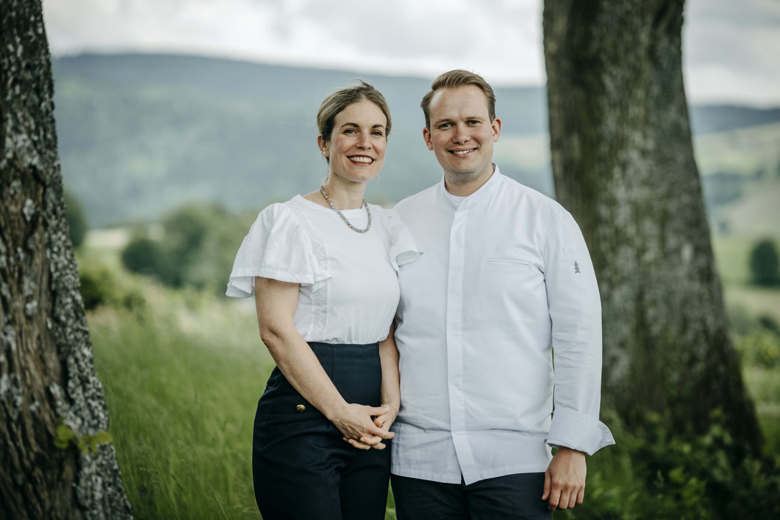 A woman and a man stand smiling outdoors between two trees, with green hills and cloudy skies in the background. She wears a white blouse and dark skirt; he wears a white chef’s jacket.