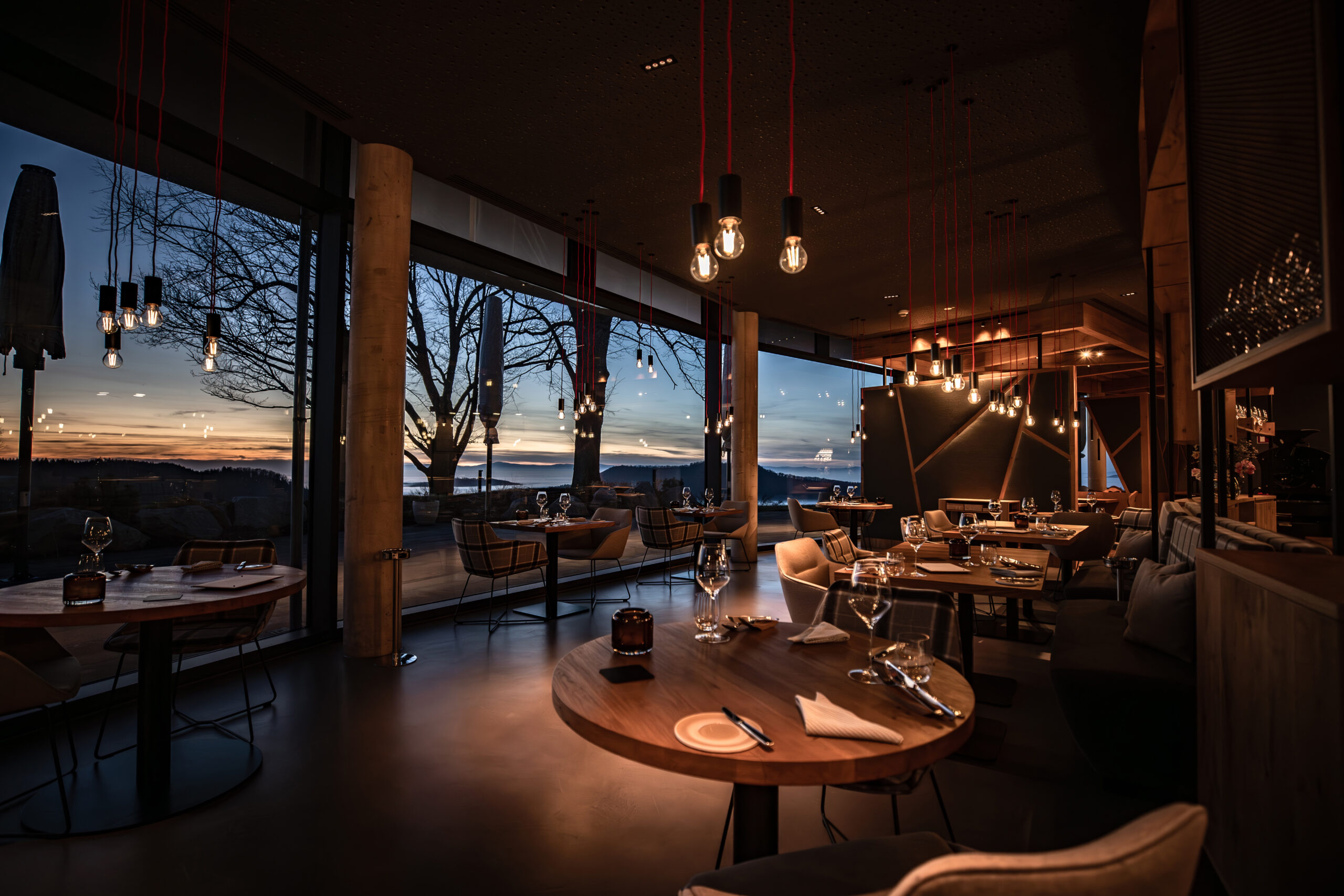 A stylish, modern restaurant interior at sunset, with round wooden tables set for dining, large windows overlooking a scenic outdoor view, and warm pendant lighting creating a cozy atmosphere.
