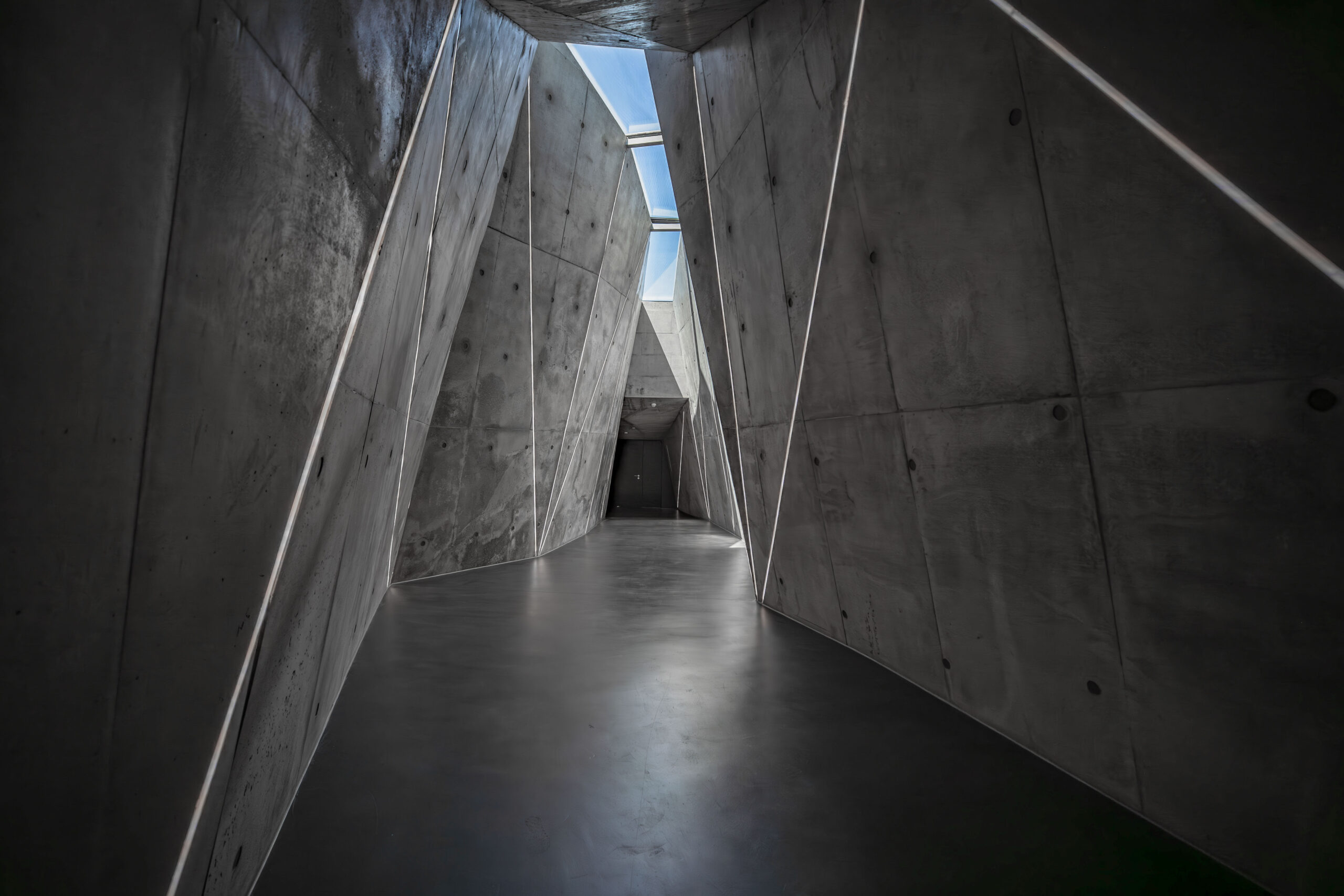 A narrow hallway with angular, concrete walls and a polished floor leads to a dark doorway. Natural light enters through a skylight above, casting shadows and highlighting the geometric architecture.