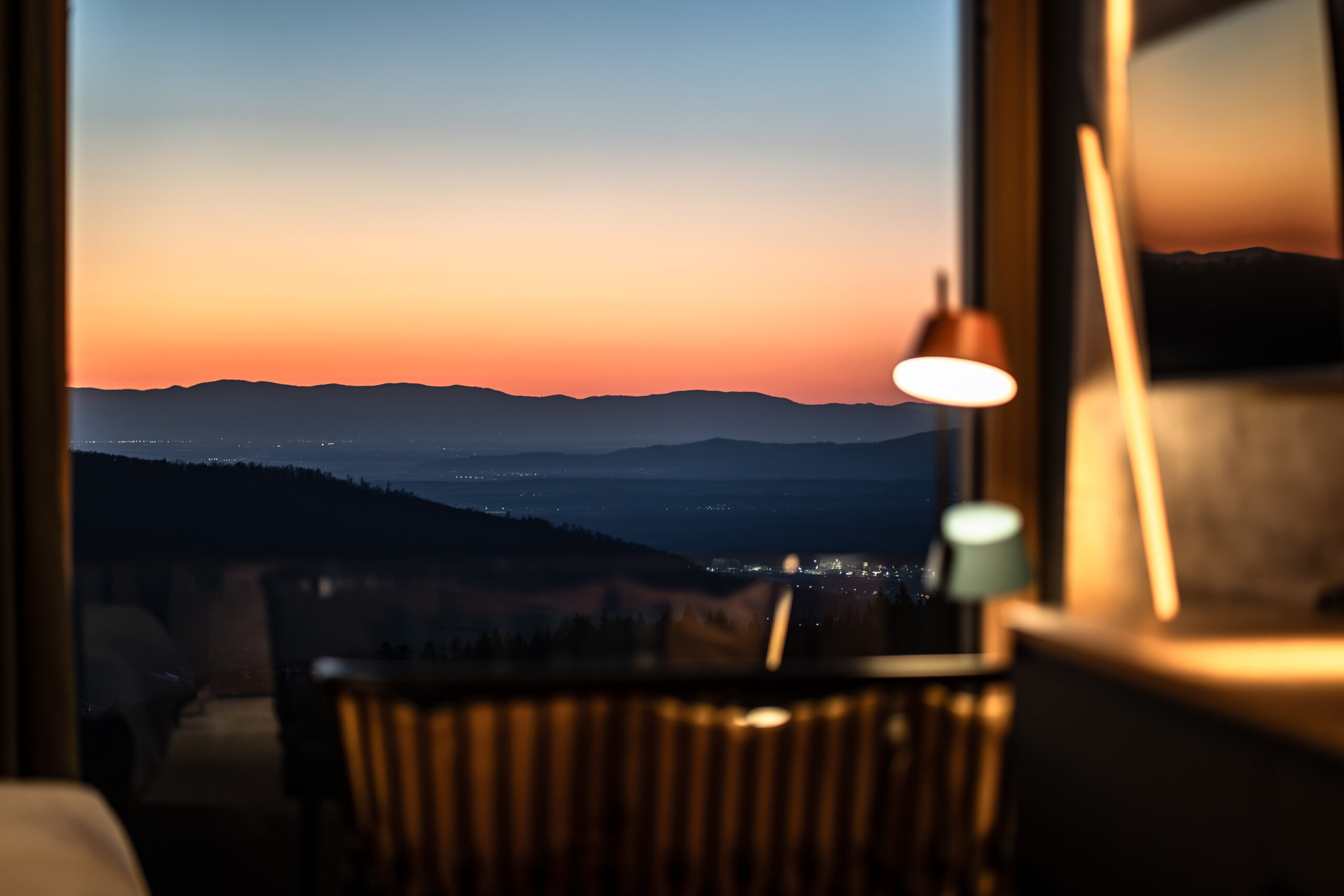 Ein gemütlicher Raum mit einem Holzstuhl und einem Schreibtisch wird von einer Lampe sanft beleuchtet, während ein großes Fenster einen atemberaubenden Blick auf die fernen Berge und den glühenden Sonnenuntergangshimmel bietet.