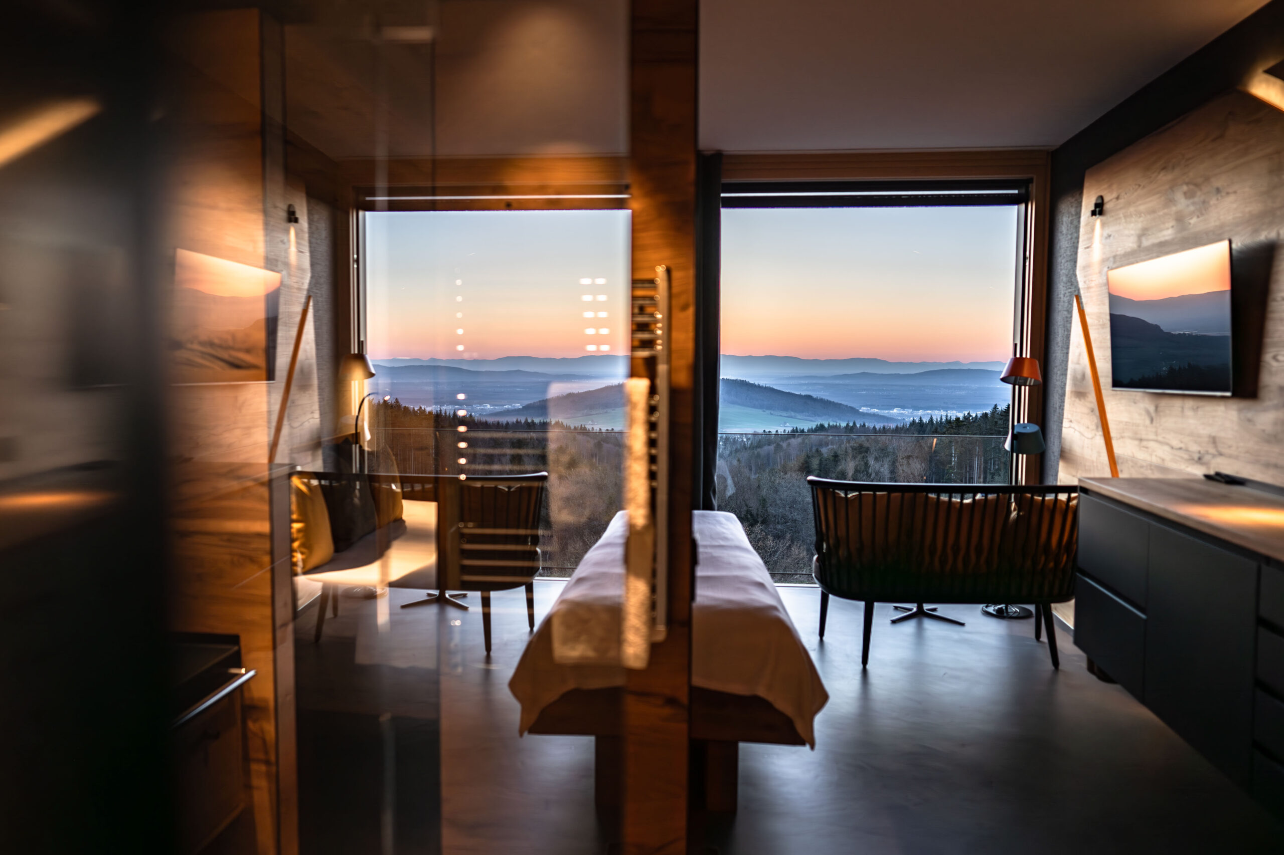 A modern hotel room with large windows overlooking a scenic mountain landscape at sunset, featuring a double bed, two chairs, a desk, and warm wooden decor.