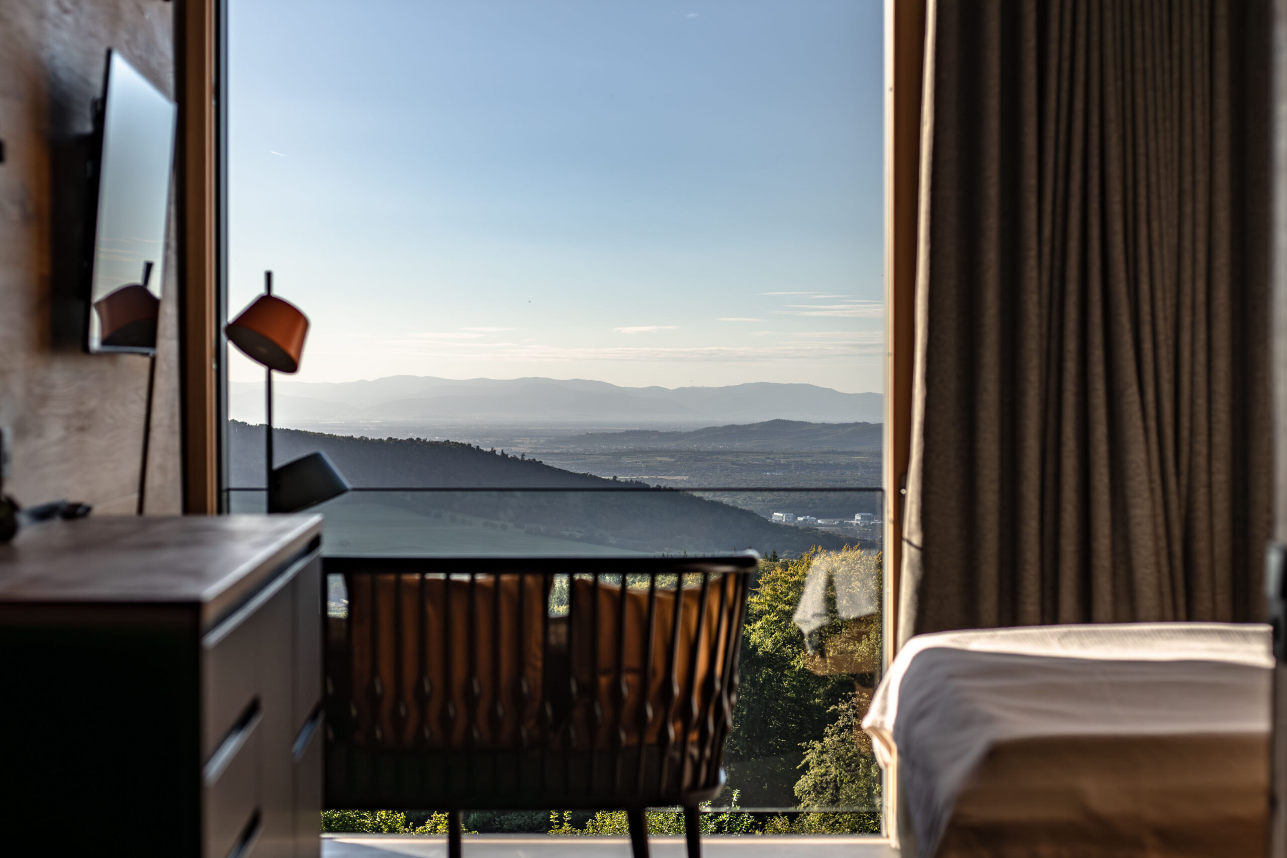 A modern room with a bed, desk, and chair faces a large window offering a scenic view of distant mountains, hills, and a clear sky.
