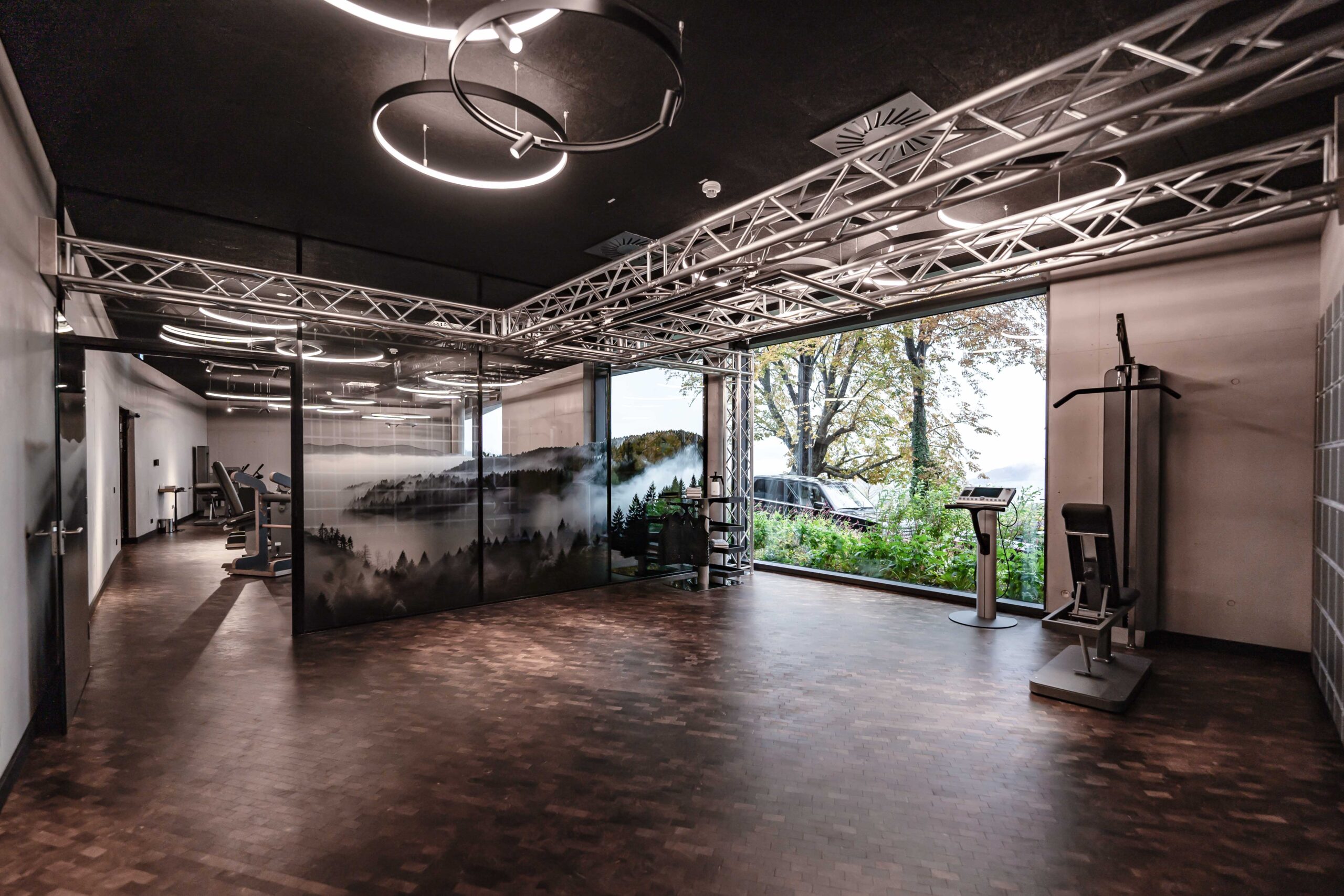 Modernes Fitnessstudio mit Holzfußboden, großen Fenstern mit Bäumen, Fitnessgeräten an den Wänden und Deckenleuchten mit einer Metallrahmenstruktur über dem Kopf. An einer Wand ist eine Landschaftsgrafik zu sehen.