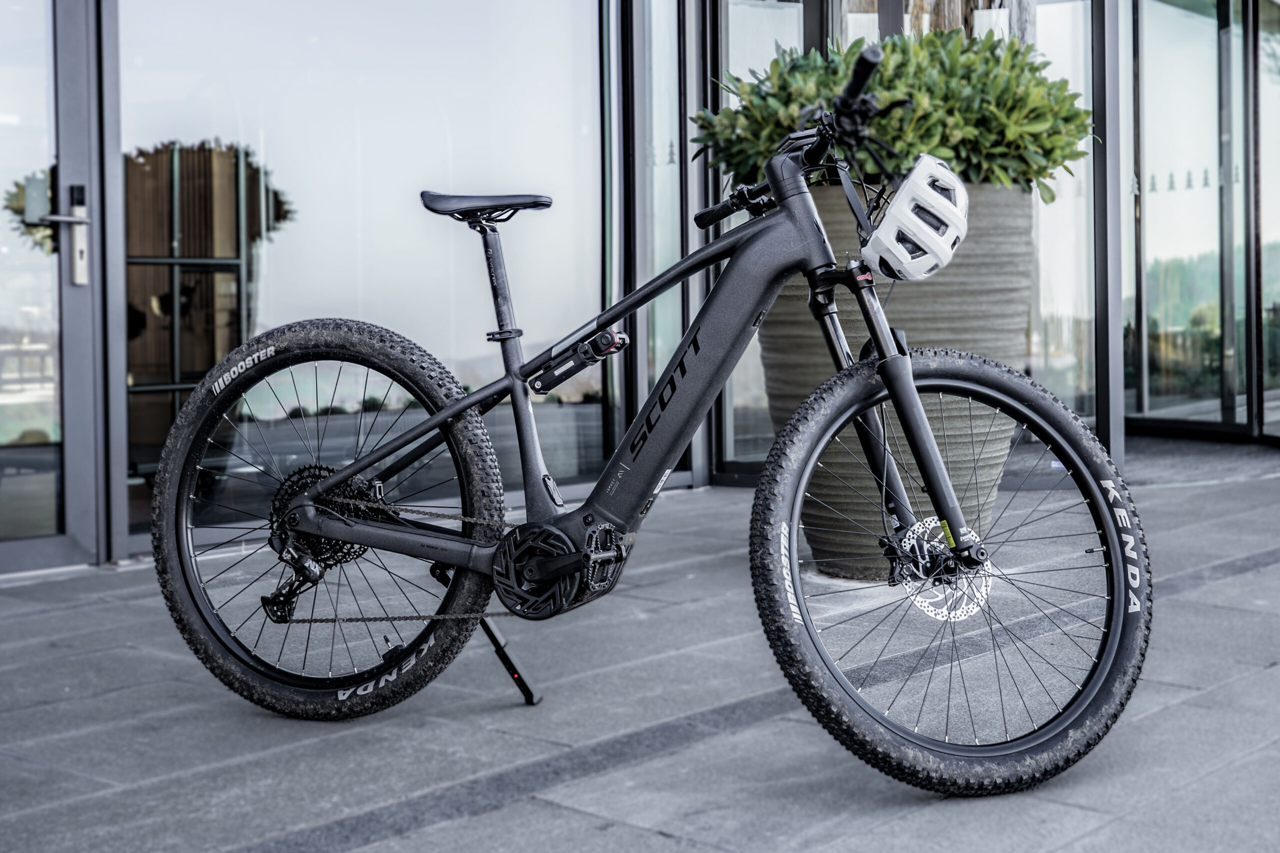 Ein schwarzes Scott-E-Mountainbike mit breiten Reifen steht auf einer grauen Steinterrasse in der Nähe einer großen Glastür und einer Topfpflanze; ein weißer Helm hängt am Lenker.