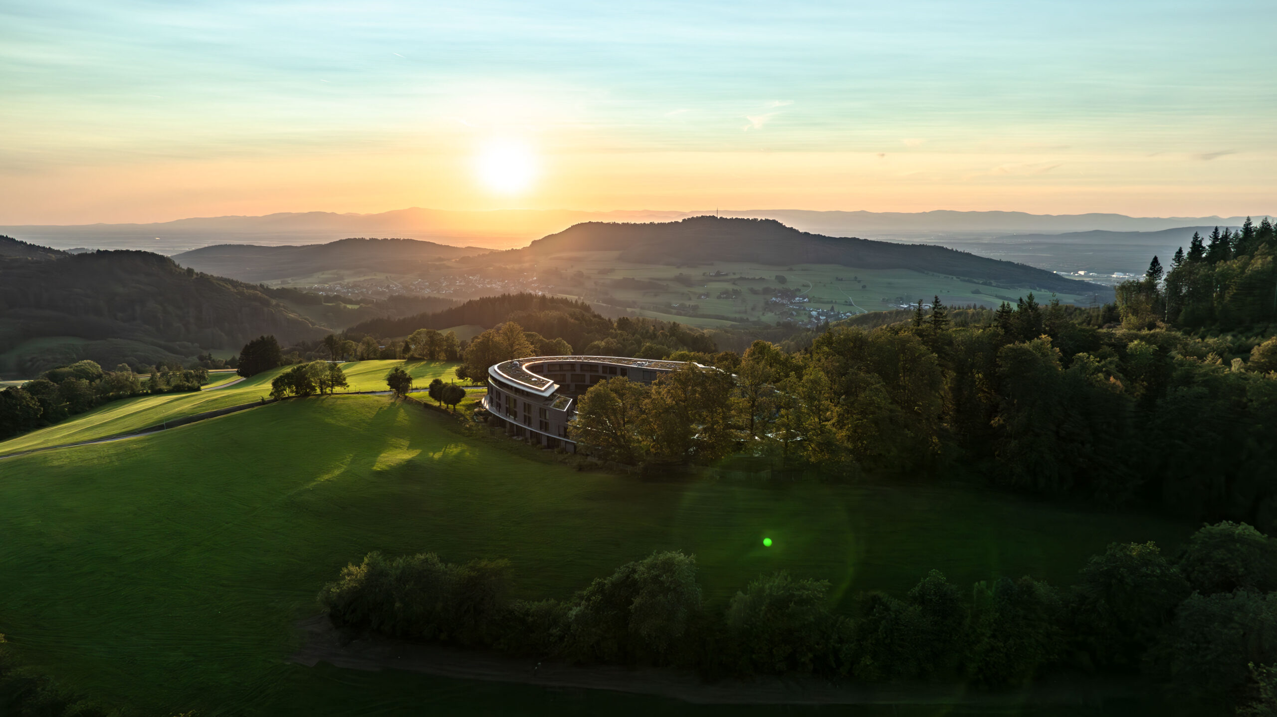 Aerial view of a modern, curved building surrounded by trees and green fields at sunset, with rolling hills and distant mountains under a colorful sky.
