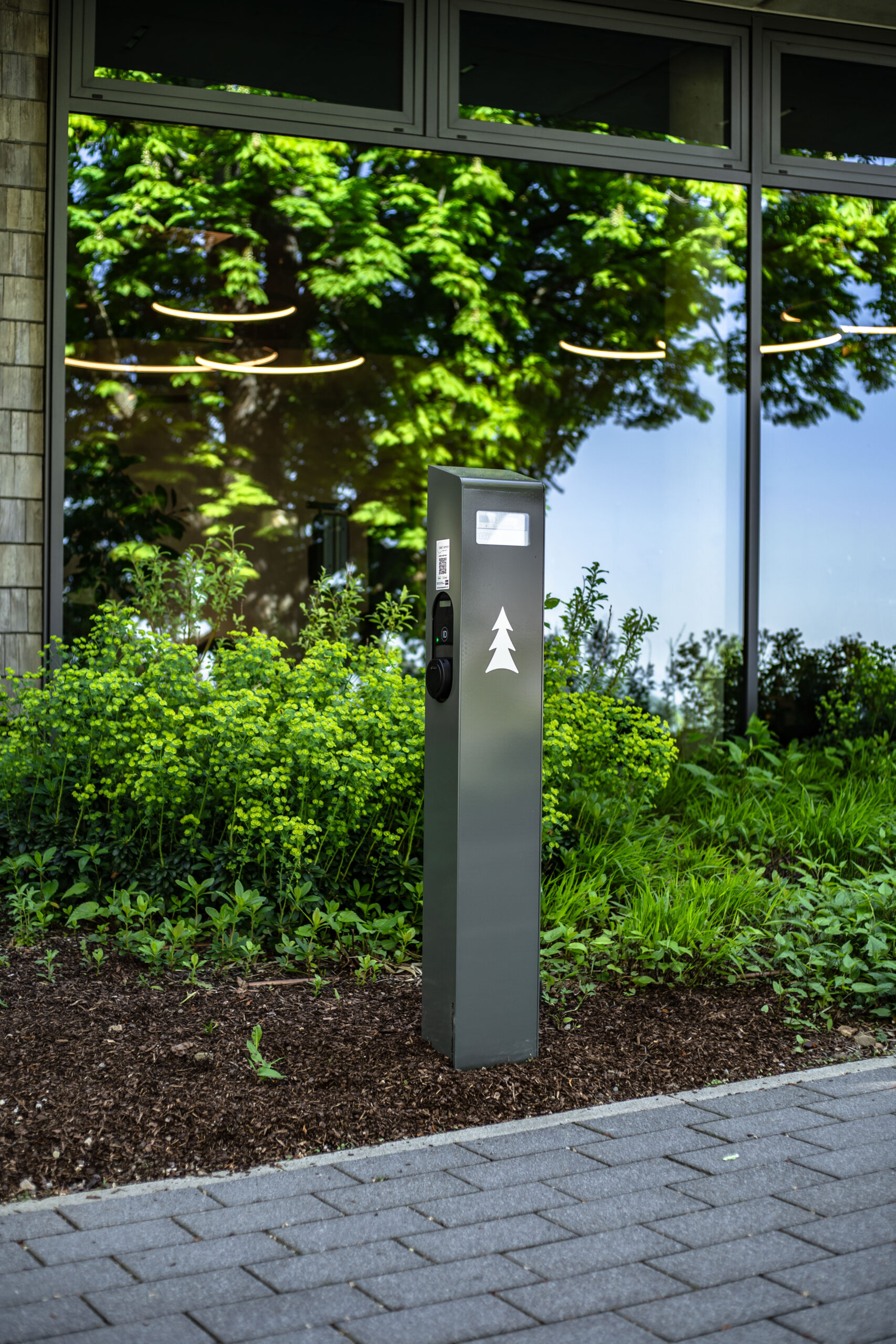 A gray electric vehicle charging station with a white tree symbol stands on a sidewalk next to green plants, in front of a glass building reflecting trees and lights.