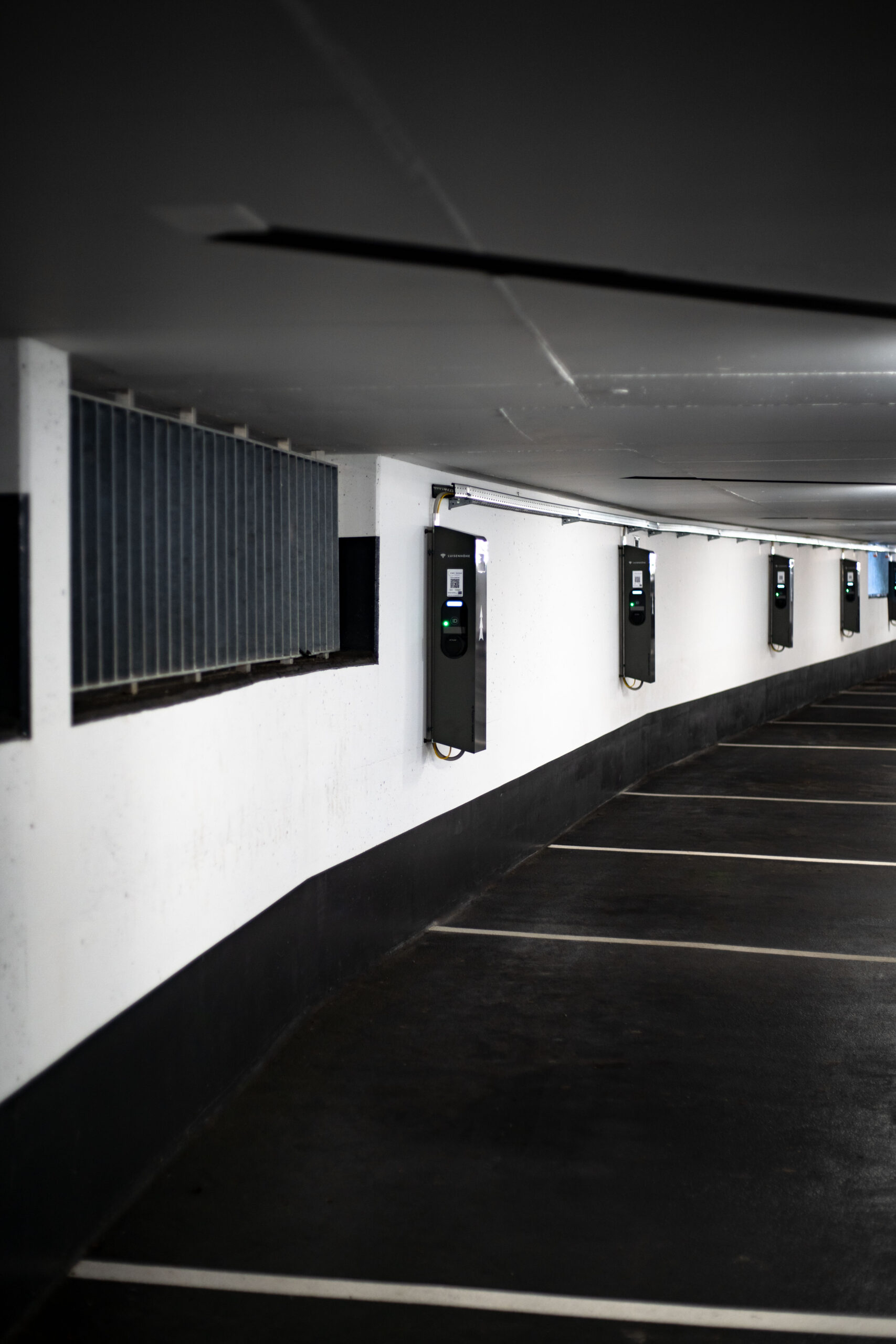 A row of empty parking spaces in an indoor garage, each space equipped with a wall-mounted electric vehicle charging station. The area is clean and dimly lit.